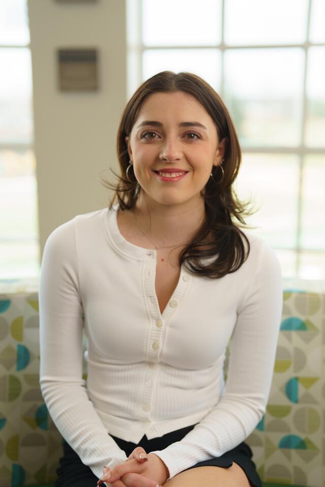 A female student with brown hair and a white top sits on a colorful couch in front of a window.