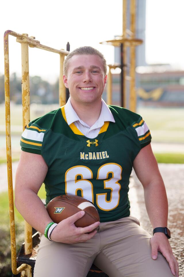 A male student wearing a football jersey sits and holds a football.