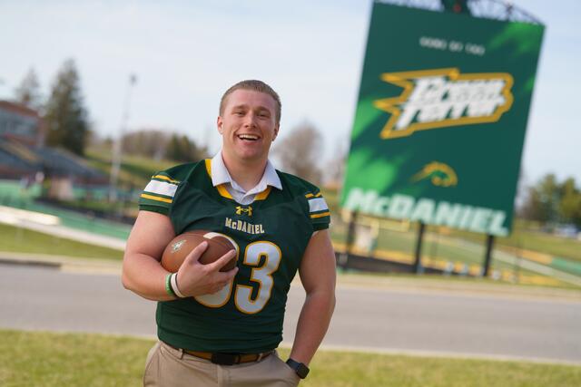 student wearing a football jersey and holding a football with a large Green Terror McDaniel scoreboard sign behind him and and the stadium in the background