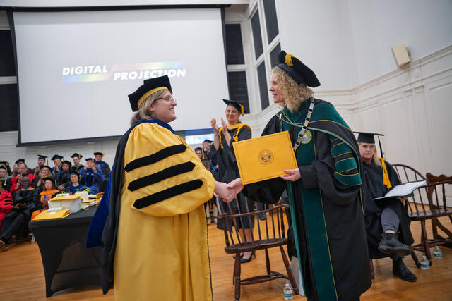 2 People in graduation regalia shake hands while holding a yellow envelope