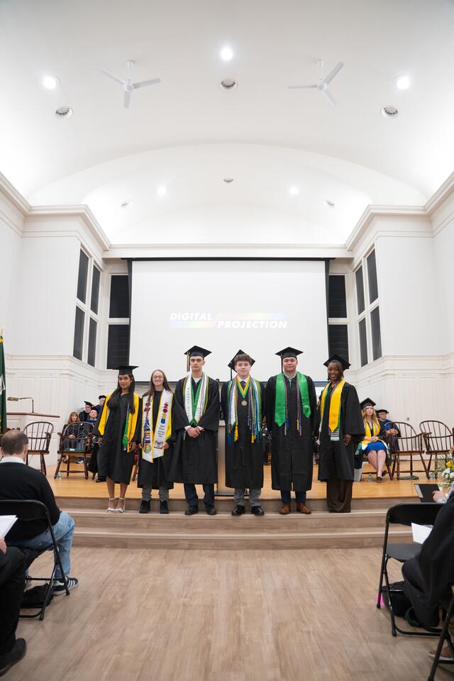Students in graduation regalia pose on stage