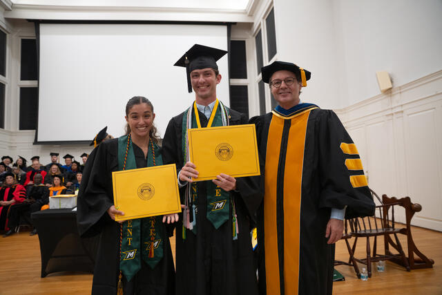 Students in graduation regalia poses with Provost Flavius Lilly, also in graduation regalia