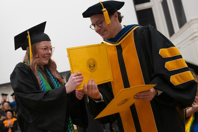 2 People in graduation regalia shake hands while holding a yellow envelope