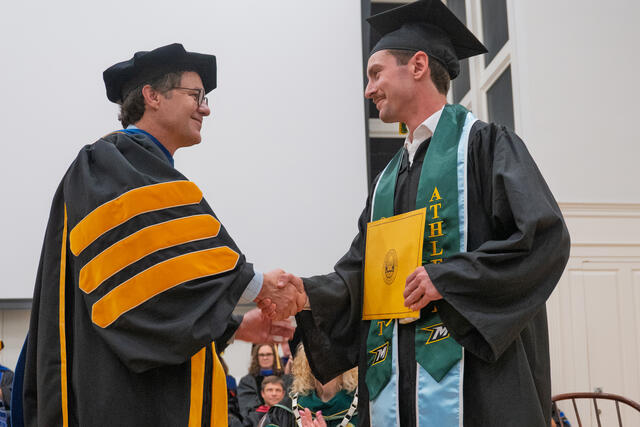 2 People in graduation regalia shake hands while holding a yellow envelope