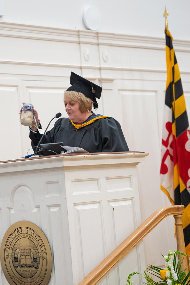 speaker at a podium, wearing graduation regalia, holding up a jar of rice