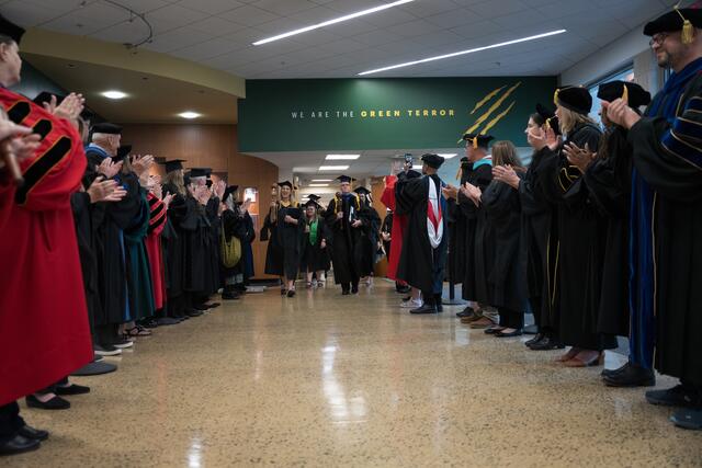 Faculty in doctoral regalia line both sides of a campus hallway and applaud as graduates process through, with a "We Are the Green Terror" sign visible on the wall.