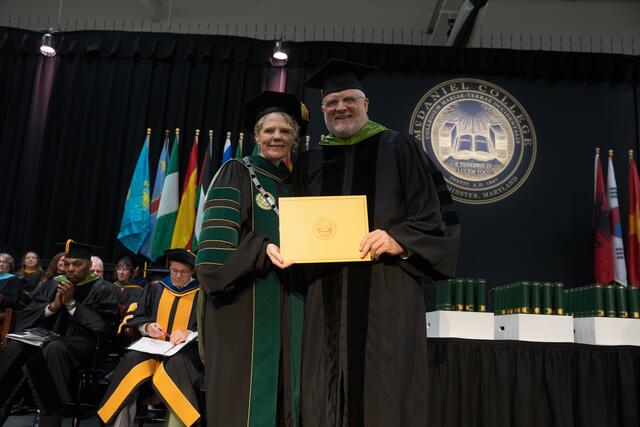 McDaniel's president and a graduate in a green stole hold a gold diploma cover together on stage, smiling, with the college seal and international flags in the background.