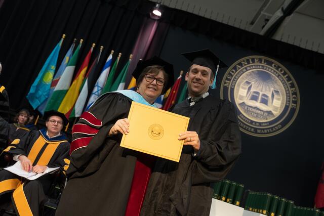 Dean Mazer, dressed in red and blue doctoral regalia, and a graduate in cap and gown hold a gold diploma cover together on stage, with the college seal visible behind them.