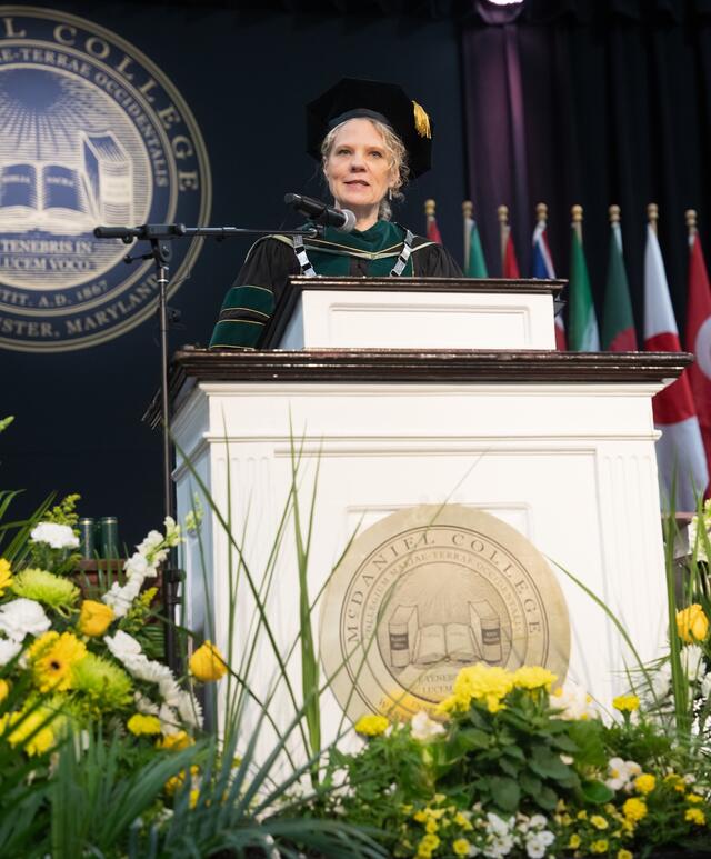 McDaniel's president speaks at the commencement podium wearing green doctoral regalia and a ceremonial chain, with the college seal and floral arrangements visible in the foreground.
