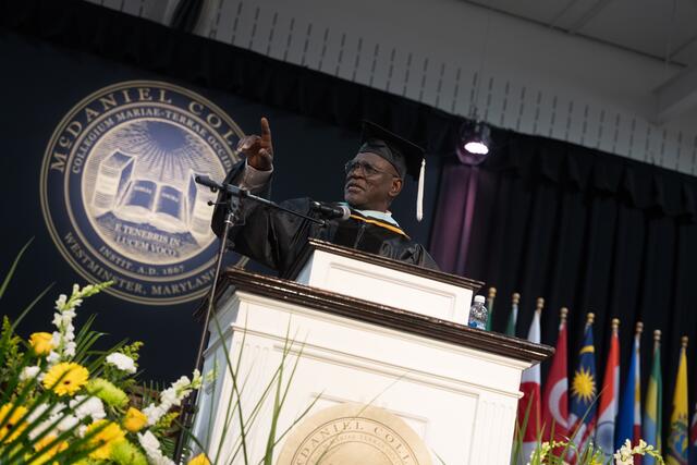 A commencement speaker in cap and gown gestures emphatically at the McDaniel College podium, with the college seal and international flags filling the background.