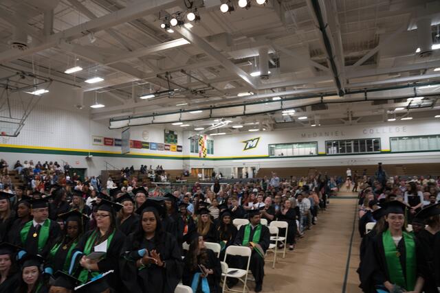 A wide view of the McDaniel graduate commencement ceremony shows graduates in green stoles seated in the gymnasium, with family and guests filling the bleachers and the McDaniel College logo visible on the wall.
