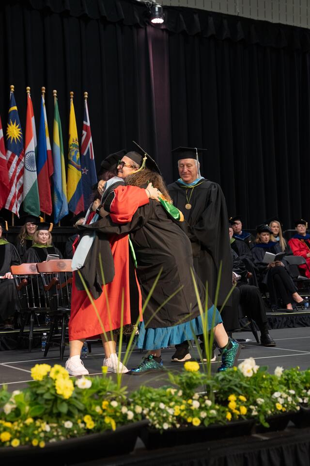 A graduate in cap and gown embraces a faculty member in red doctoral regalia on the commencement stage, while another faculty member looks on with a smile.