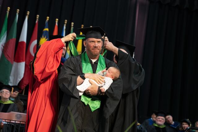 A graduate in cap and gown holds an infant while two faculty members place a doctoral hood over their head on the commencement stage, with international flags in the background.