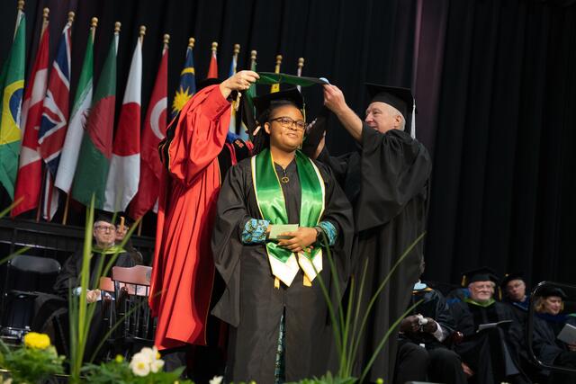 Two faculty members lower a doctoral hood over a graduate's head on the commencement stage as the graduate holds a diploma and smiles, with international flags in the background.