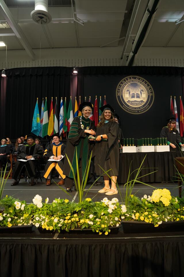 McDaniel's president shakes hands with a graduate while presenting a diploma scroll on stage, with the college seal, international flags, and floral arrangements visible in the background.