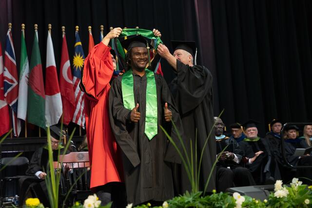 Two faculty members lift a doctoral hood over a smiling graduate's head on the commencement stage, with the graduate giving a thumbs up and international flags in the background.