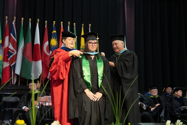  Two faculty members lower a doctoral hood over a graduate's head on the commencement stage, with international flags and faculty seated in the background.