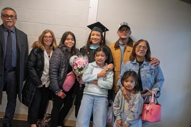  A graduate in cap and gown holds a bouquet of flowers while posing with family members of multiple generations after the commencement ceremony.