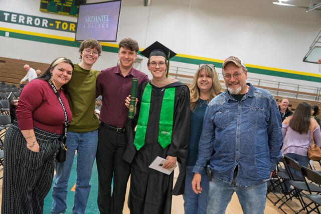 A graduate in cap, gown, and green McDaniel College stole holds a diploma scroll and poses with family members in the gymnasium after the commencement ceremony, with a McDaniel College screen visible in the background.
