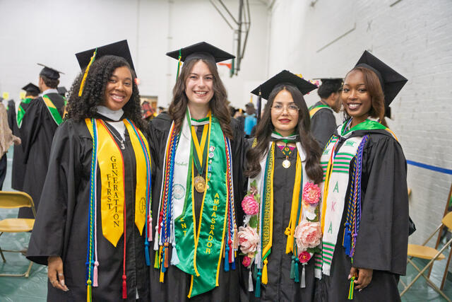 Four graduates in caps and gowns smile together, wearing various honor stoles and cords including a "First Generation" stole and a "Student Ambassador" sash.