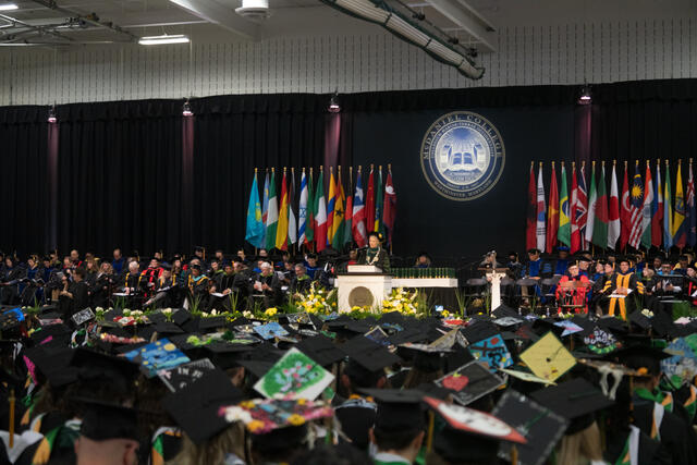A wide shot of McDaniel College commencement shows rows of graduates in decorated caps facing a speaker at the podium, with the college seal visible behind the stage.