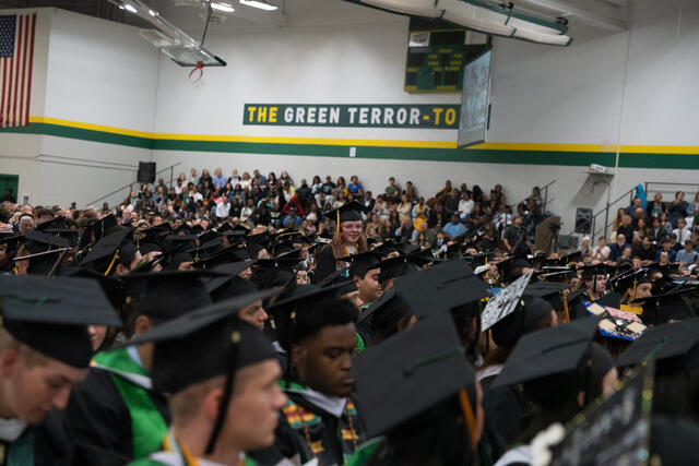 Rows of graduates in caps and gowns fill the gymnasium during commencement, with a "The Green Terror" sign visible on the wall above.