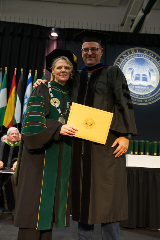 McDaniel College's president, wearing ceremonial chain and green doctoral regalia, poses with a graduate holding a gold diploma cover in front of the college seal.