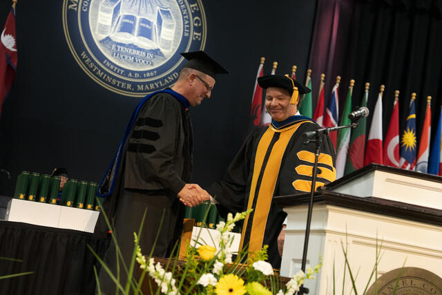 Two people in doctoral regalia shake hands on stage at the commencement podium, with international flags in the background.