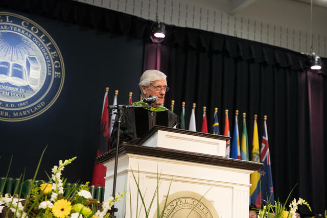 A speaker in green-trimmed doctoral regalia addresses the crowd from the McDaniel College commencement podium, surrounded by international flags.