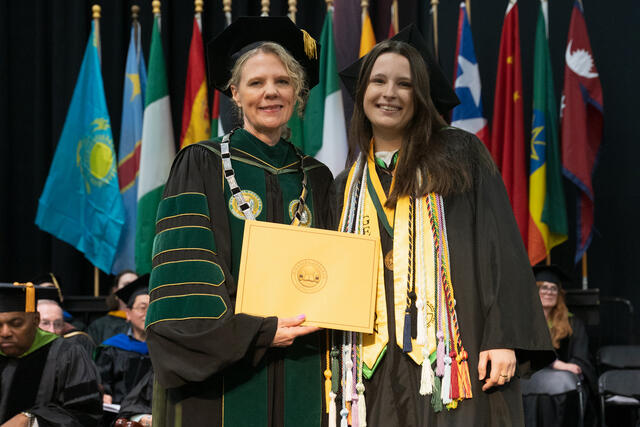 McDaniel's president and a graduate hold a gold diploma cover together on stage, smiling, with international flags in the background.
