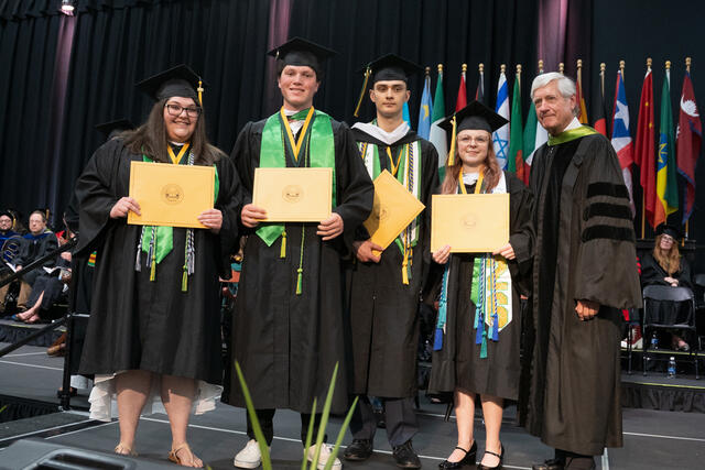 Four graduates holding gold diploma covers pose on stage with a trustee Bruce Preston '75, with flowers and international flags visible in the background.