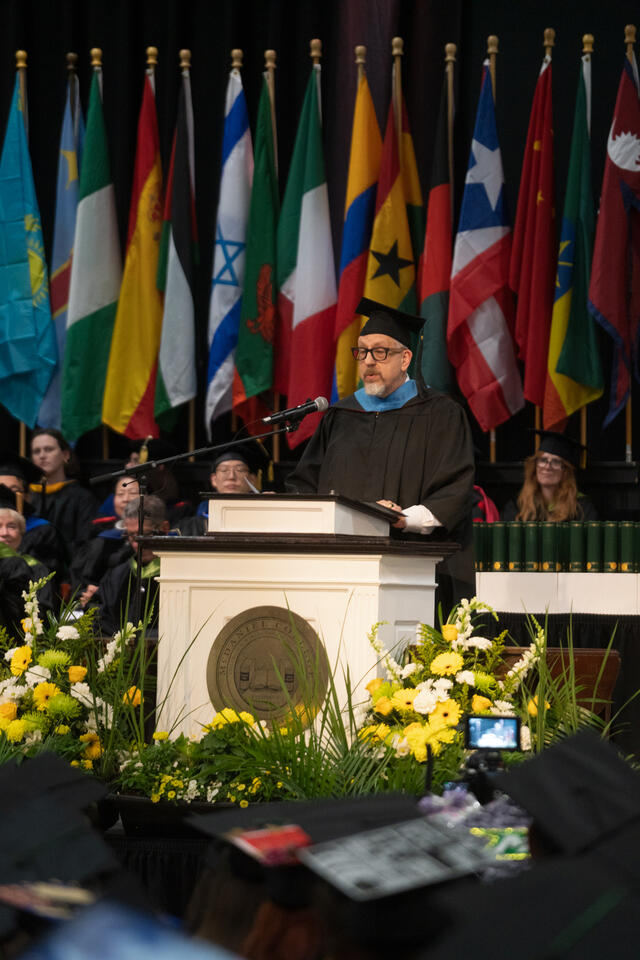 Trustee Bob Pick, dressed in blue-trimmed doctoral regalia speaks at the McDaniel College commencement podium, with international flags filling the background.