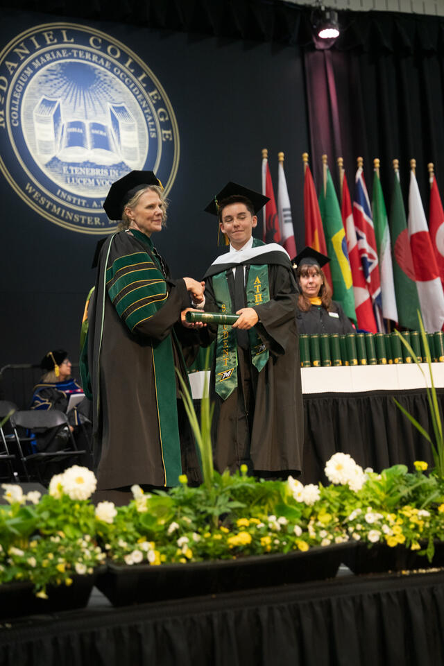 McDaniel's president hands a diploma scroll to a graduate wearing a green "Student Athlete" stole, with the college seal and international flags behind them.