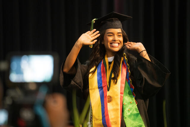 A graduate in cap and gown waves and grins at the crowd while crossing the commencement stage, wearing a gold "Global Fellow" stole and honor medal.