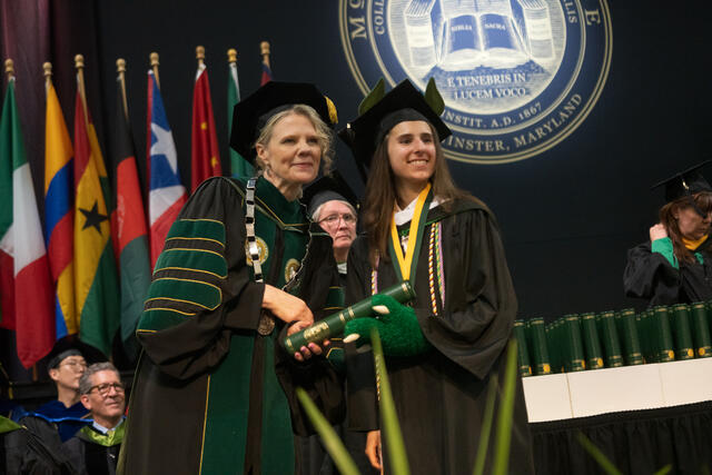 McDaniel's president and a smiling graduate hold a diploma scroll together on stage, with the college seal and international flags in the background.