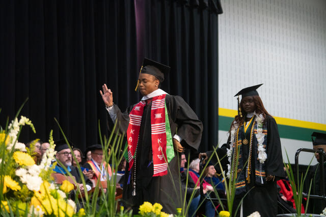 Two graduates cross the commencement stage, one wearing a red Kappa Alpha Psi stole and raising a hand, the other wearing a floral lei and kente stole.