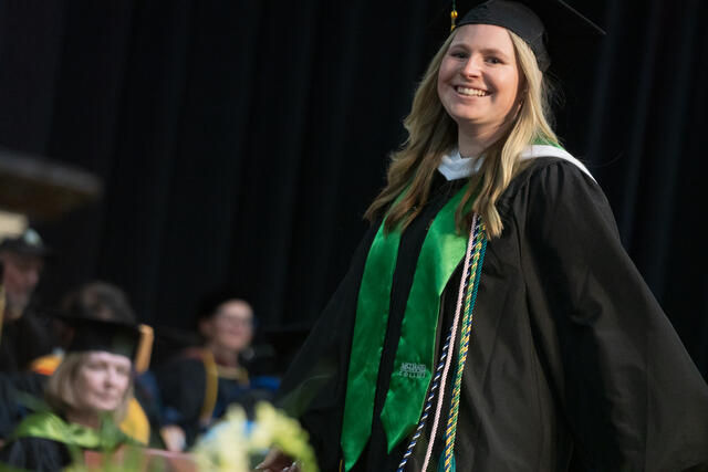A smiling graduate in cap, gown, and green McDaniel College stole walks across the commencement stage, with faculty seated in the blurred background.