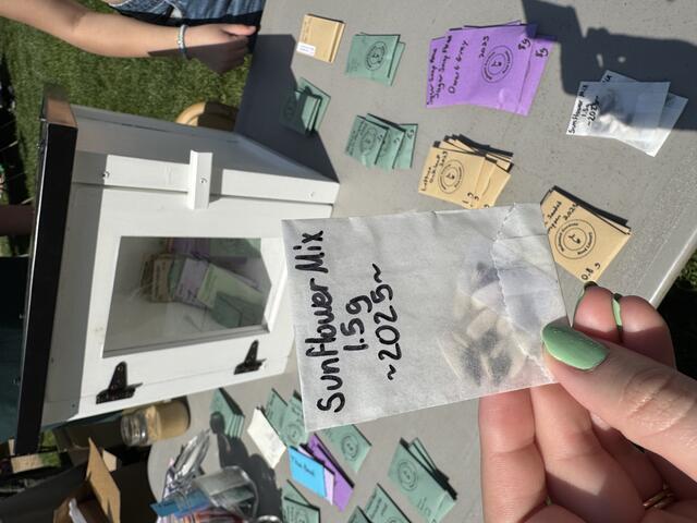A hand holds up a packet of seeds labeled "Sunflower Mix" in front of the seed library.