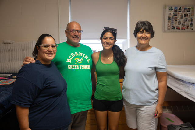 A family of four people standing in a room, smiling. One person is wearing a green shirt with "McDaniel Green Terror" printed on it. The room appears to be in a residence hall with bunk beds and a bulletin board in the background.