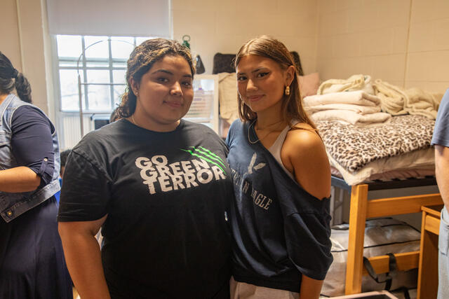 Two students standing in a dorm room, one wearing a "Go Green Terror" shirt and the other an American Eagle top. A bed with patterned bedding is in the background.