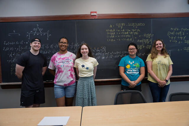 A group of students stands in front of a chalkboard filled with mathematic equations.