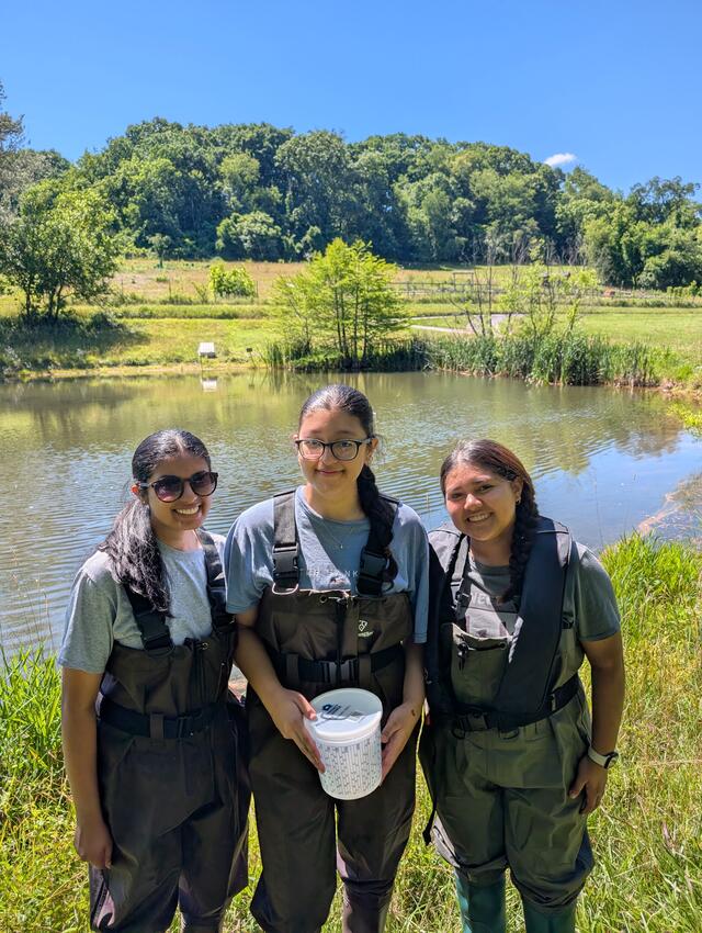 Three students stand by a pond and wear wading gear.