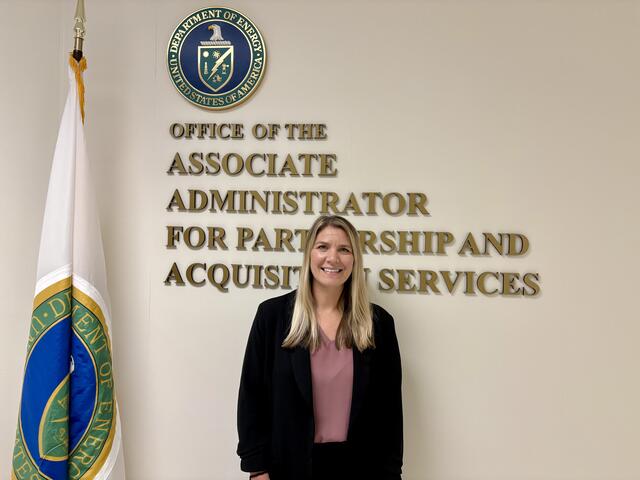 A blonde woman in professional wear stands in front of a sign for the Office of the Associate Administrator for Partnership and Acquisition Services at the United States Department of Energy.