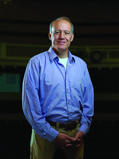 Professor Richard Sautter stands in a blue button up in the campus theatre.