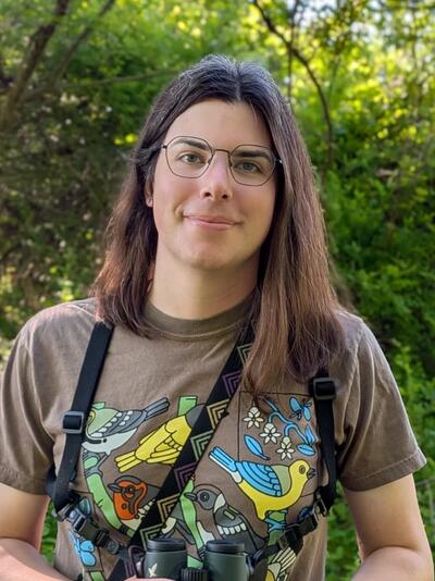 person with brown hair stands in front of trees