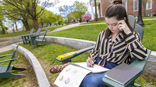 Student drawing in sketchpad while sitting in outdoor chair on campus.