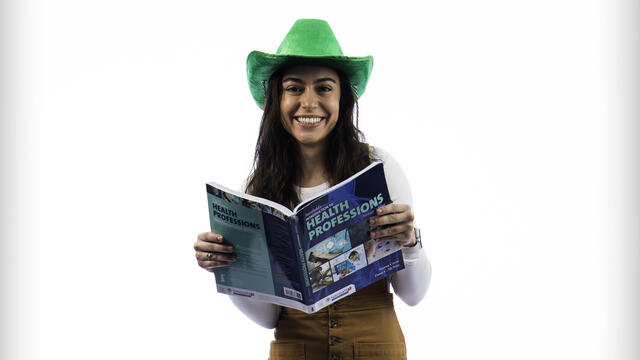 Giulia Pires holds a textbook on health professions open while wearing a green cowboy hat.