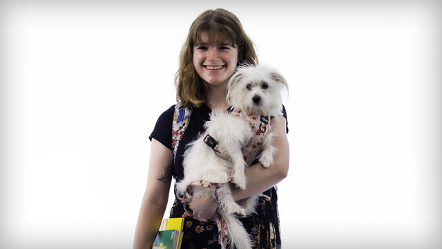 Micaela Champion poses while holding a textbook and a small white dog.