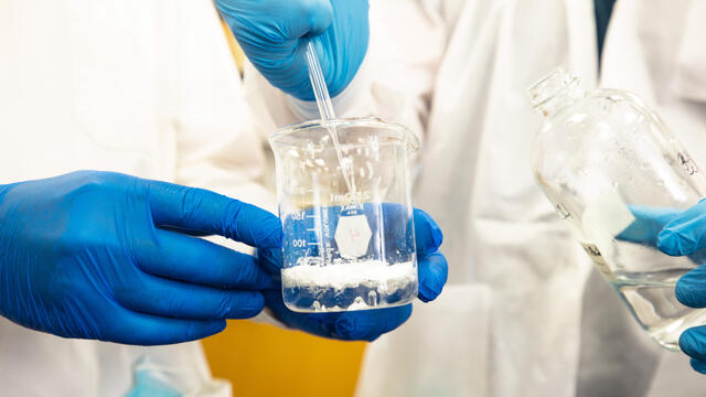 Close-up of hands in latex gloves handling a beaker and pipette with solution in them.