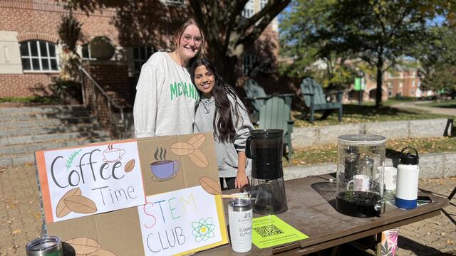 Two students stand at a table with a sign for STEM Club and coffee carafes.